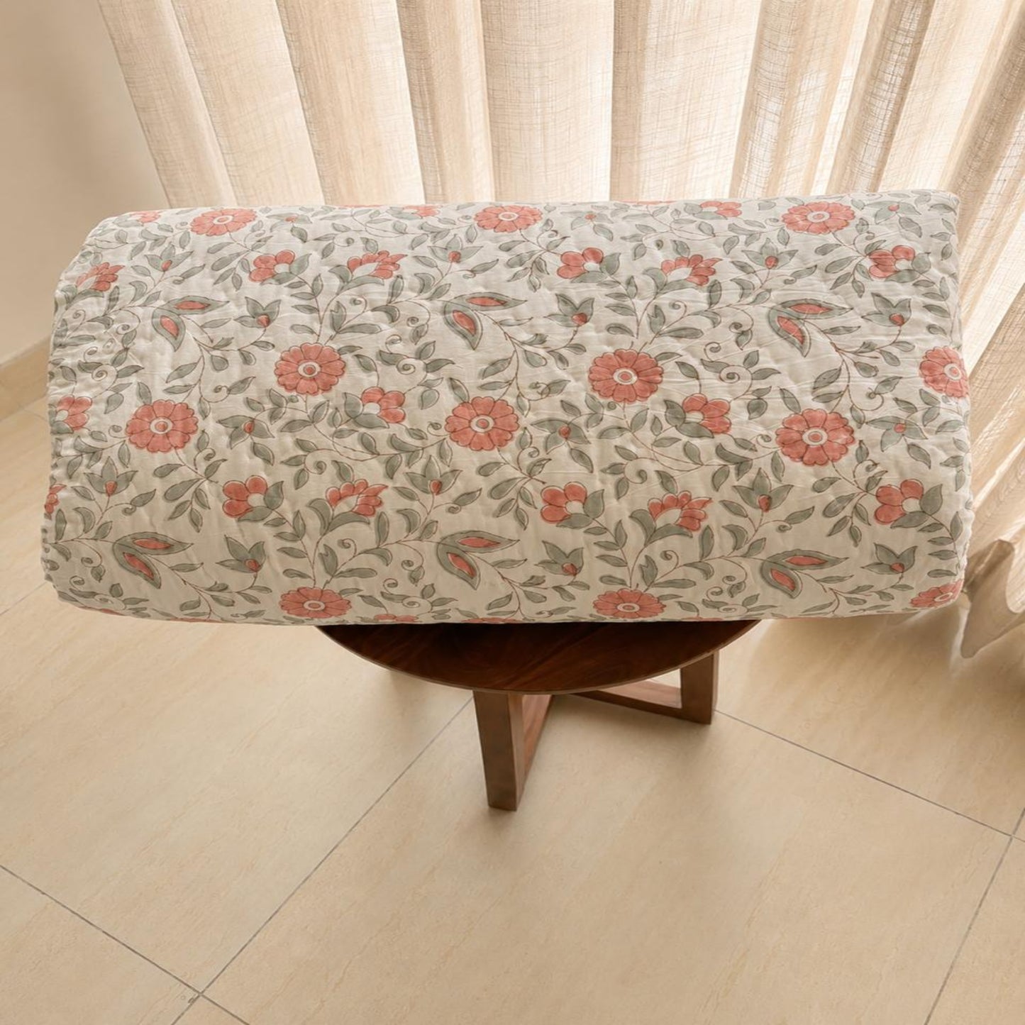 A floral pattern in coral pink and muted green on a cream background, draped over a small wooden stool in front of sheer curtains.