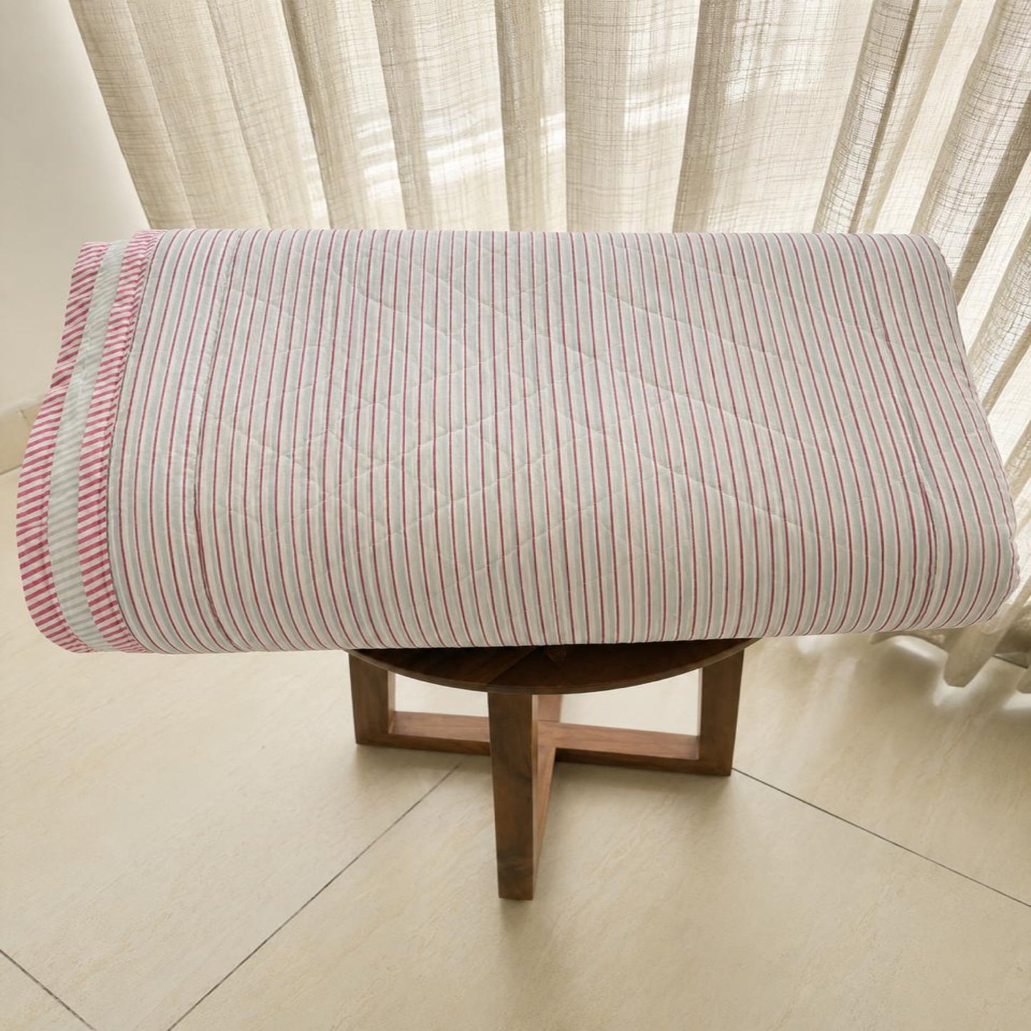 A rolled Jaipur Razai/Comforter/Quilt with thin red and white vertical stripes and a matching striped edge, displayed on a small wooden stool in front of sheer curtains.
