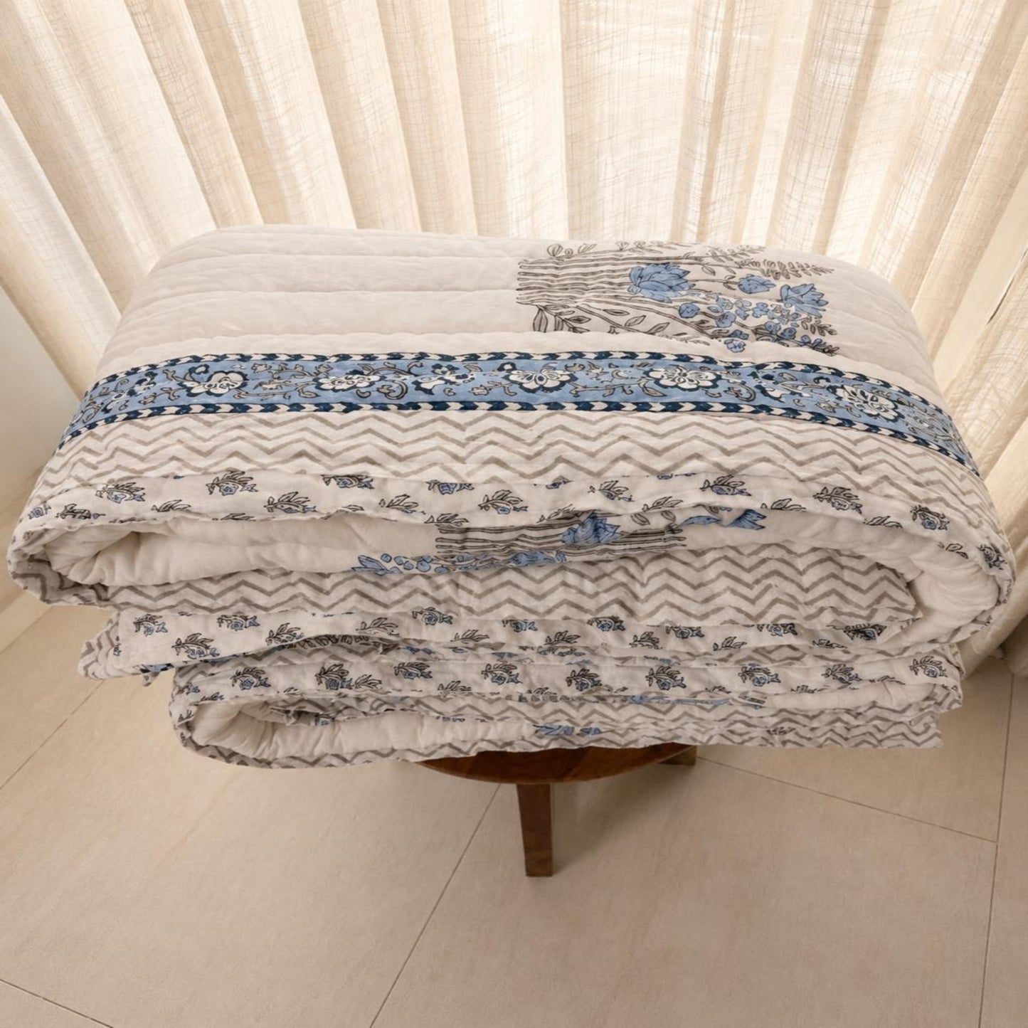 A folded Jaipuri Block Print Razai with a blue and gray patterned border and floral motifs, displayed on a small wooden stool in front of cream-colored curtains.