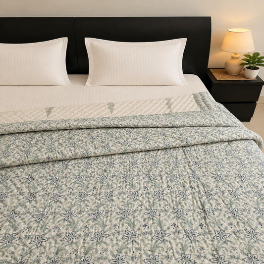 A neatly made bed with white pillows and sheets, featuring a folded light sage green Jaipuri Block Print Razai/Quilt/Comforter with a blue floral pattern. The bed is set against a dark wood headboard, with a matching dark nightstand on the right holding a lit lamp and a small potted plant.