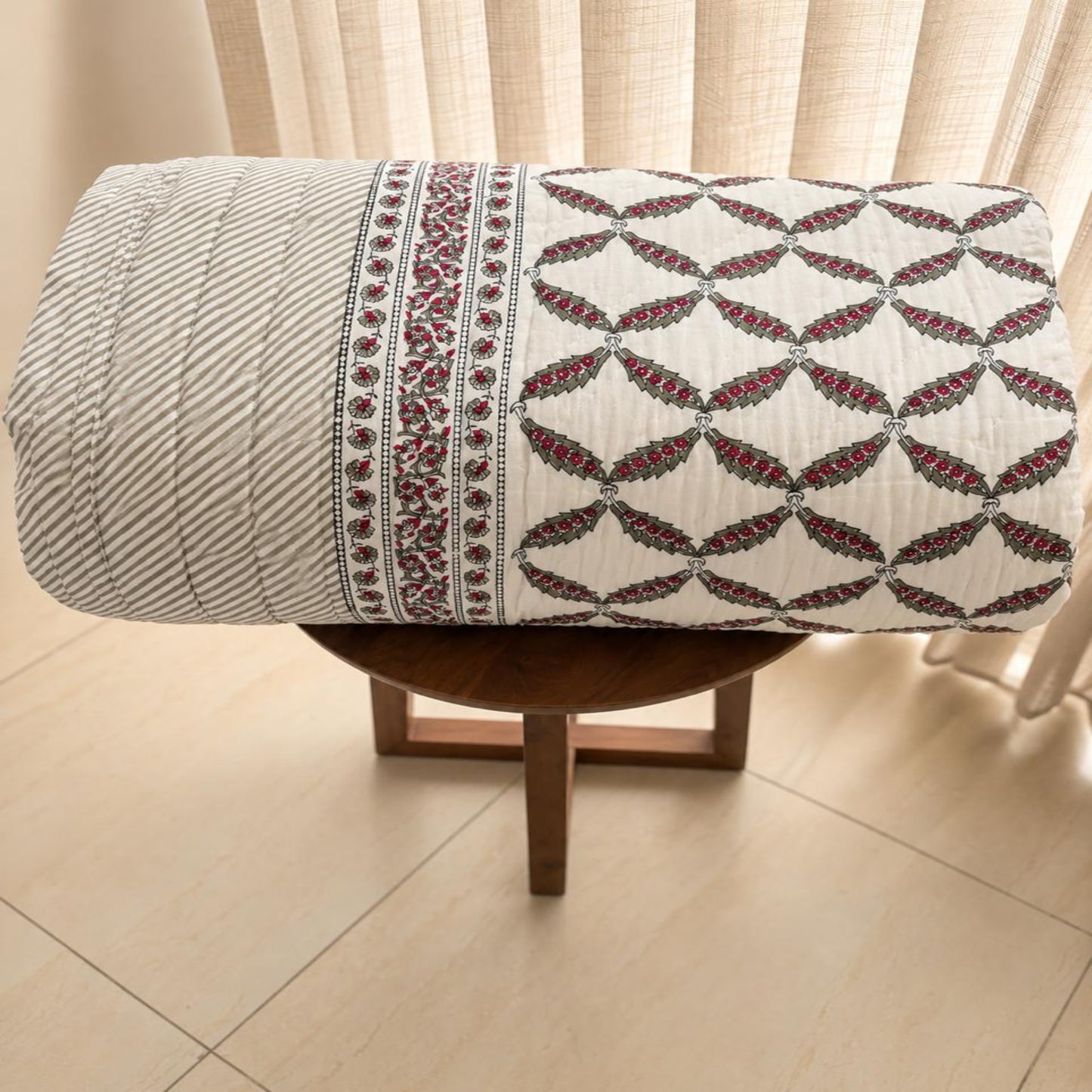 A rolled-up beautiful Jaipuri Razai with intricate burgundy and green block-printed patterns, displayed on a small wooden stool in front of beige curtains.