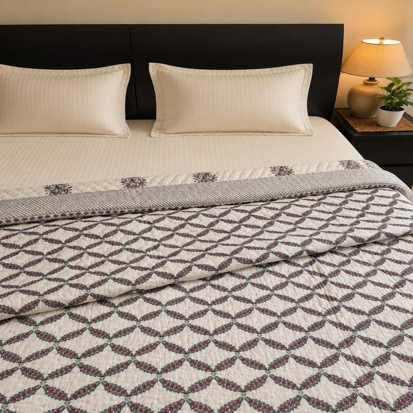A neatly made bed with two cream pillows, a cream fitted sheet, and a beautiful patterned Razai/Comforter/Quilt. The quilt has a repeating geometric leaf-like motif in gray and muted burgundy on an off-white base.