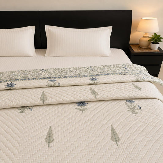 A neatly made bed with white pillows and a beautiful Jaipuri Razai/Quilt/Comforter featuring a blue floral border and tree motifs, styled in a bedroom with a dark brown headboard and bedside lamp.