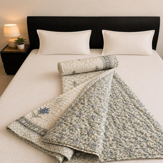 A lovely Jaipuri Razai, featuring a light off-white quilt with a blue-gray floral pattern and matching pillowcases, styled in a warm, neutral bedroom setting.
