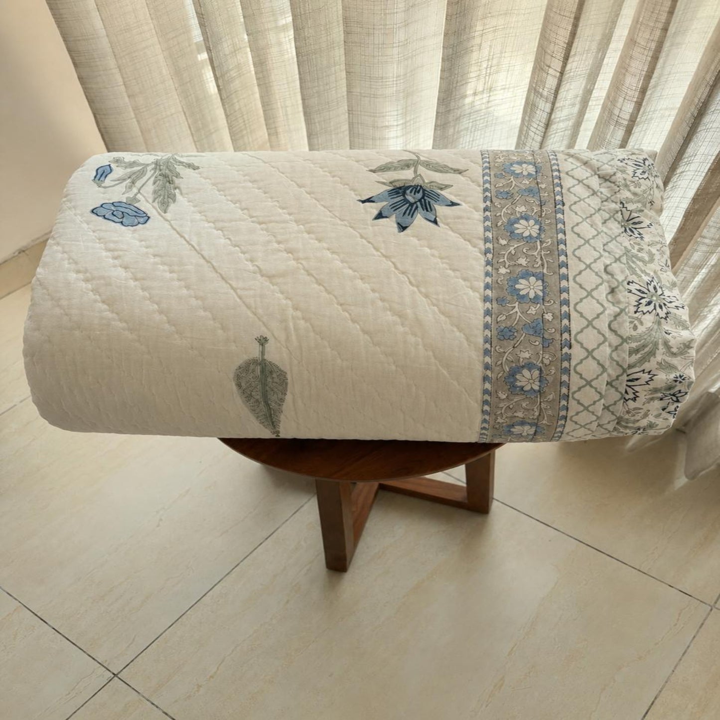 A rolled Jaipuri Razai, displayed on a small wooden stool in front of sheer curtains. The fabric is off-white/cream with blue and green botanical leaf patterns and a decorative border.