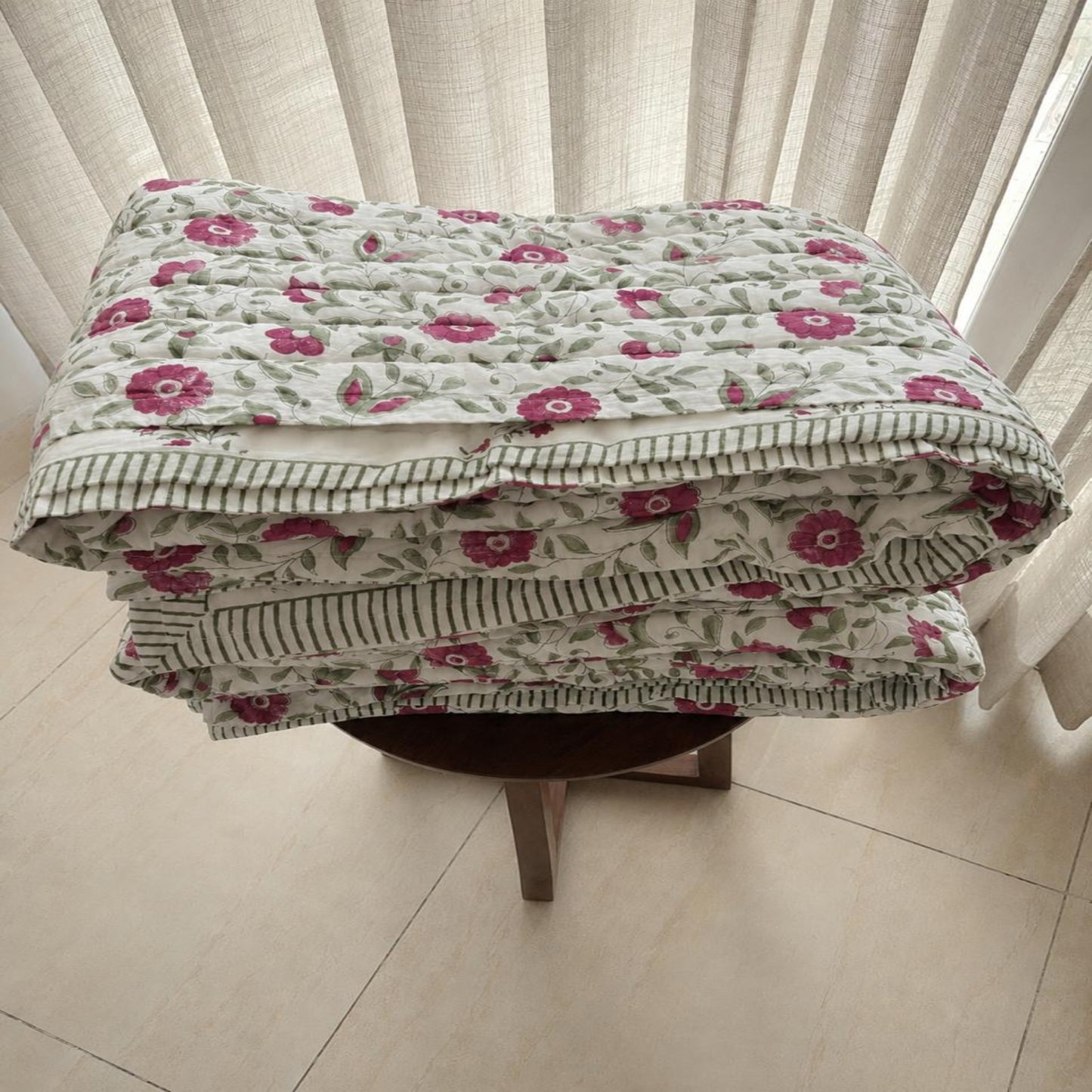 A folded Jaipuri razai with pink and green floral pattern on a light background, displayed on a small wooden stool in front of white curtains.