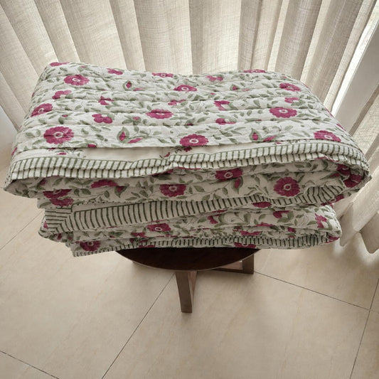 A folded Jaipuri razai with pink and green floral pattern on a light background, displayed on a small wooden stool in front of white curtains.