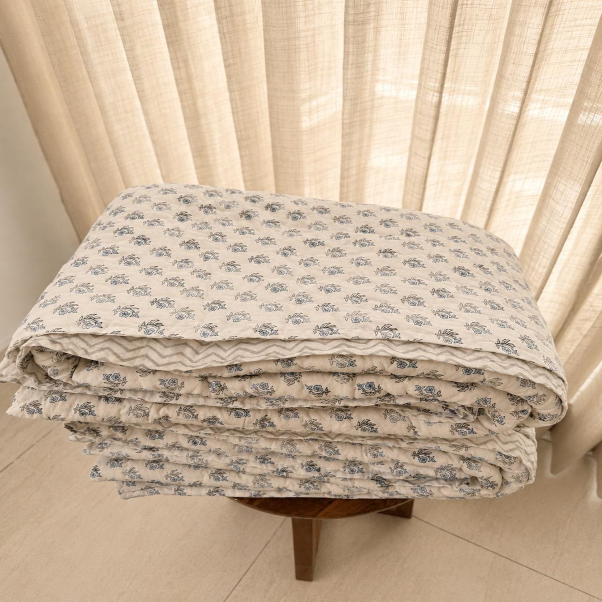 A folded beautiful Jaipuri Razai with small blue floral pattern on a white background, displayed on a wooden stool in front of beige curtains.