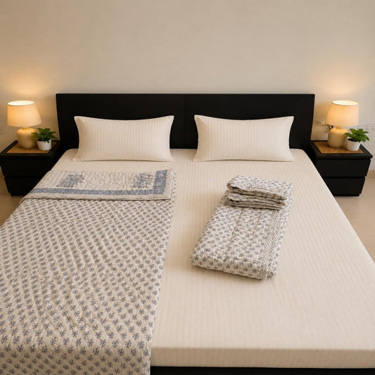 A neatly made bed in a minimalist bedroom setting, featuring a cream-colored mattress, two white pillows, a folded patterned Jaipuri Block Print Cotton Razai, and a matching folded textile stack on the right side. The bed is flanked by matching dark bedside tables with lit lamps and small potted plants.