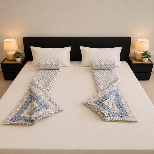 A neatly made bed with a pair of Jaipuri Razais, each folded to show a blue and white patterned design, set against a dark headboard with bedside lamps and plants.