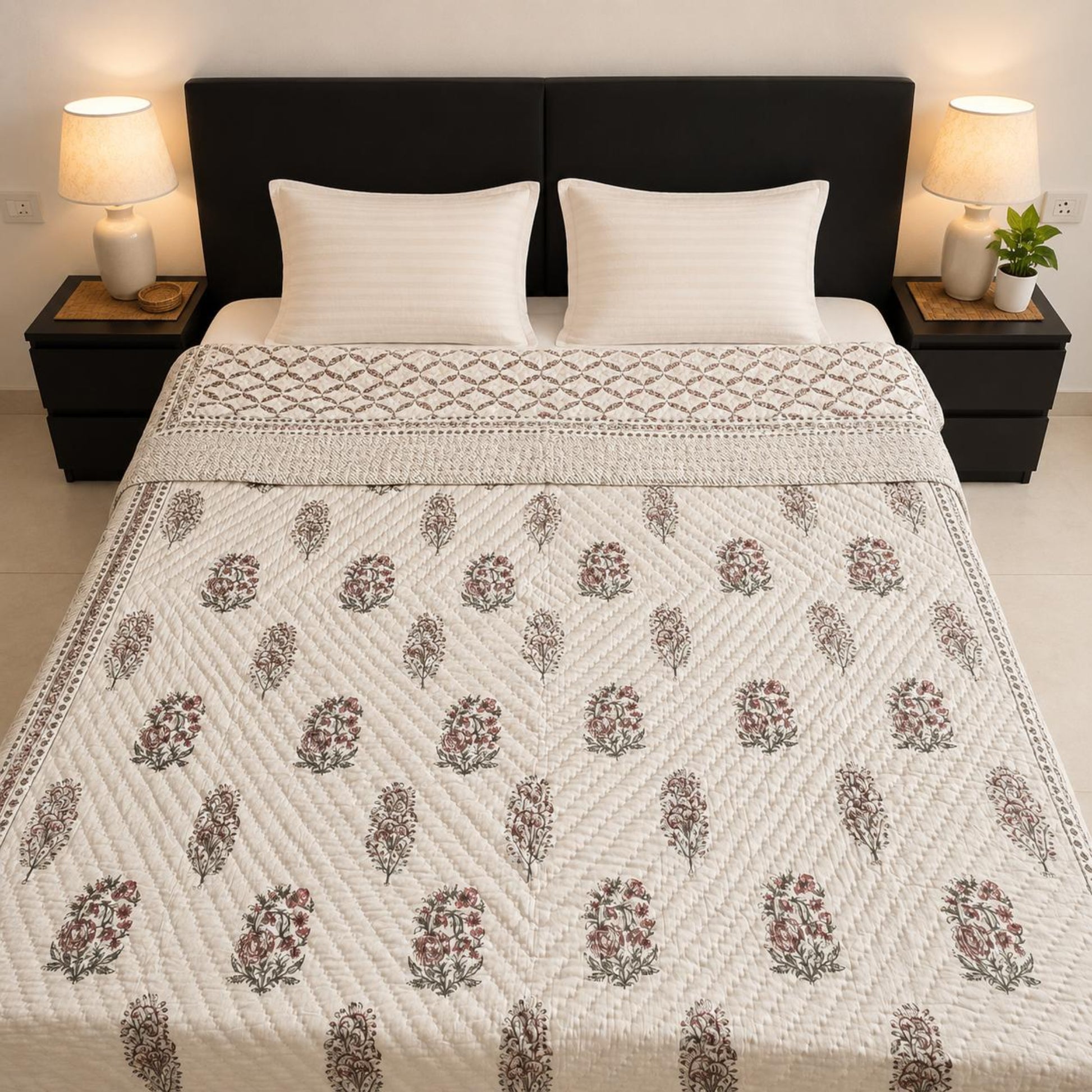 An elegant Jaipuri Razai/Quilt with a repeating floral block-print pattern in muted maroon, green, and brown on an off-white background, styled on a bed with two white pillows and dark brown nightstands.