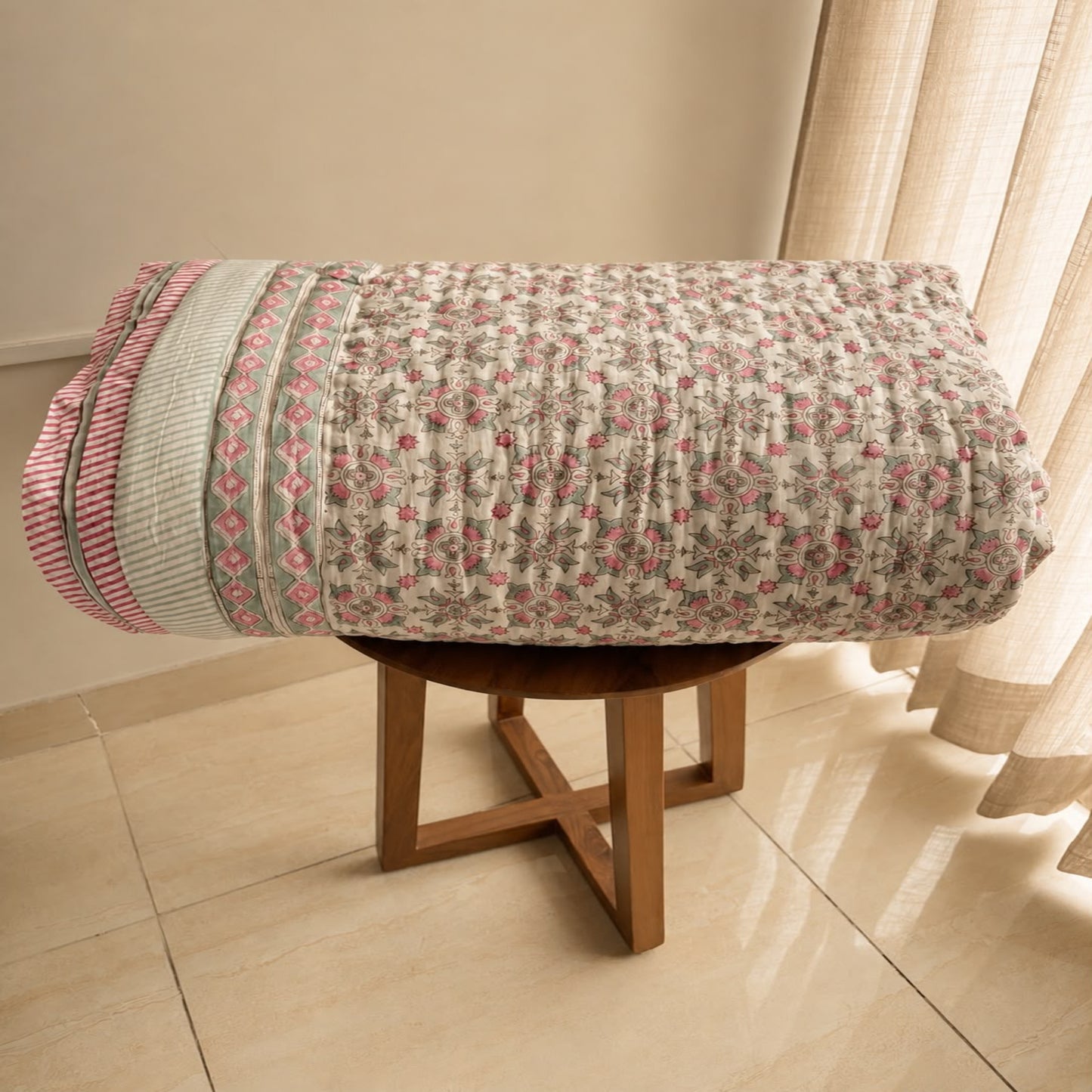 A folded Jaipuri razai/comforter. A pin floral pattern and striped border, displayed on a wooden stool in an indoor room with tiled floor and curtain.