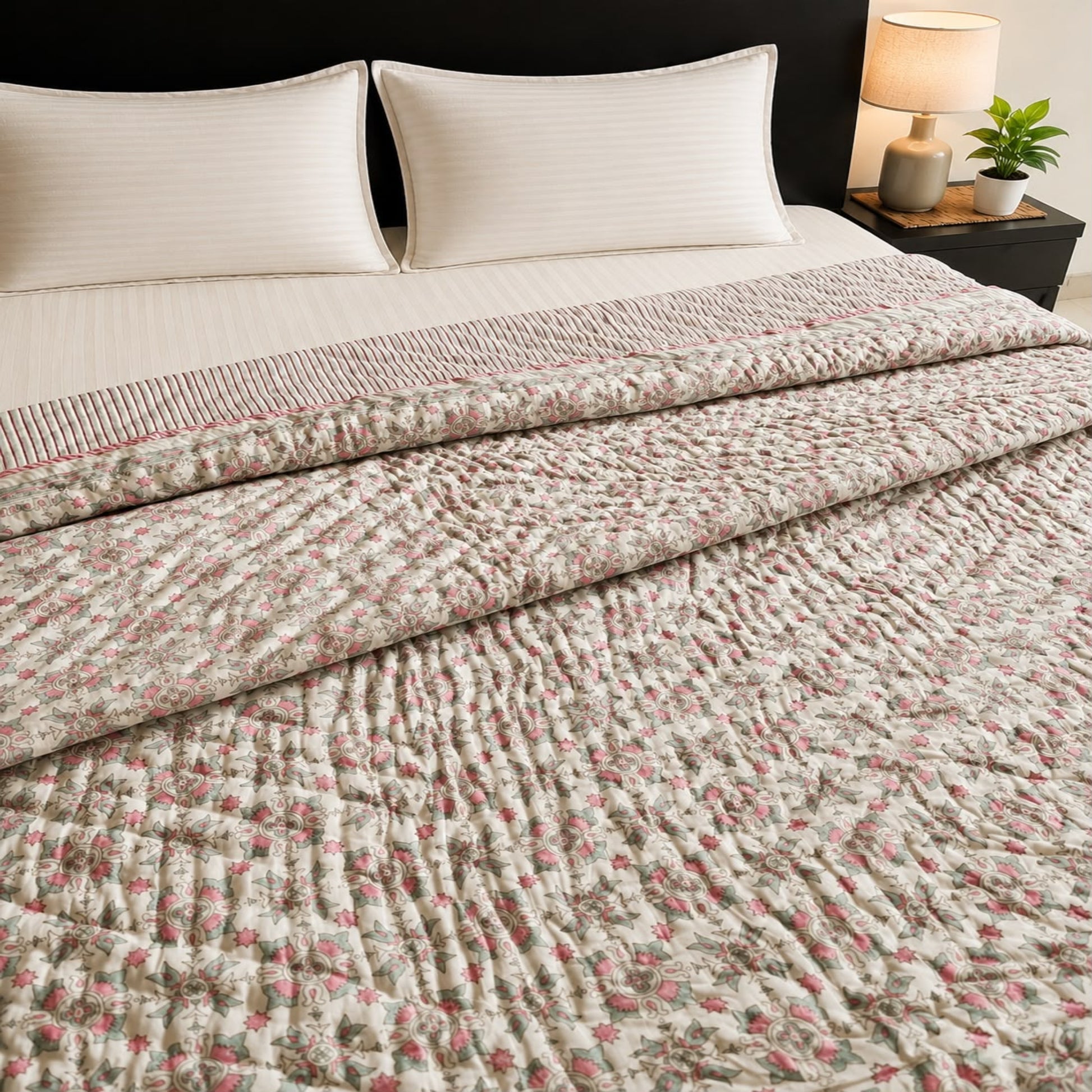 A reversible Jaipuri razai/quilt/comforter with a pink floral pattern, styled on a bed with cream pillows and a bedside lamp.
