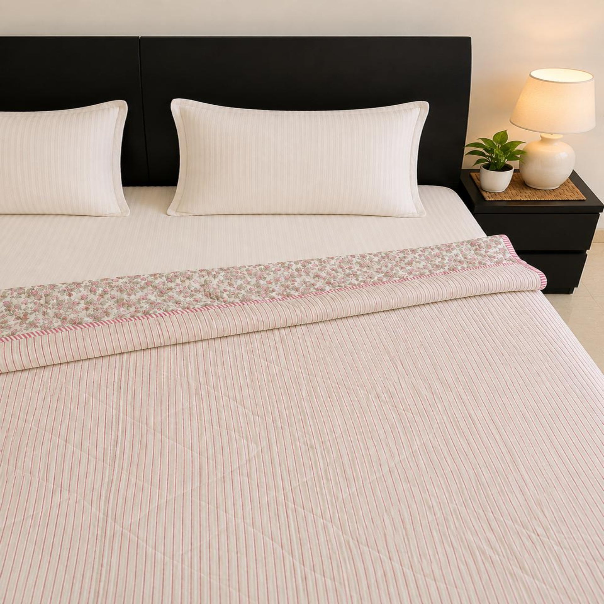 A neatly made bed with a delicate stripe patterned and pink edging Jaipuri Razai/Quilt/Comforter, two white pillows, and a dark headboard. A black bedside table holds a lit lamp and a small potted plant.