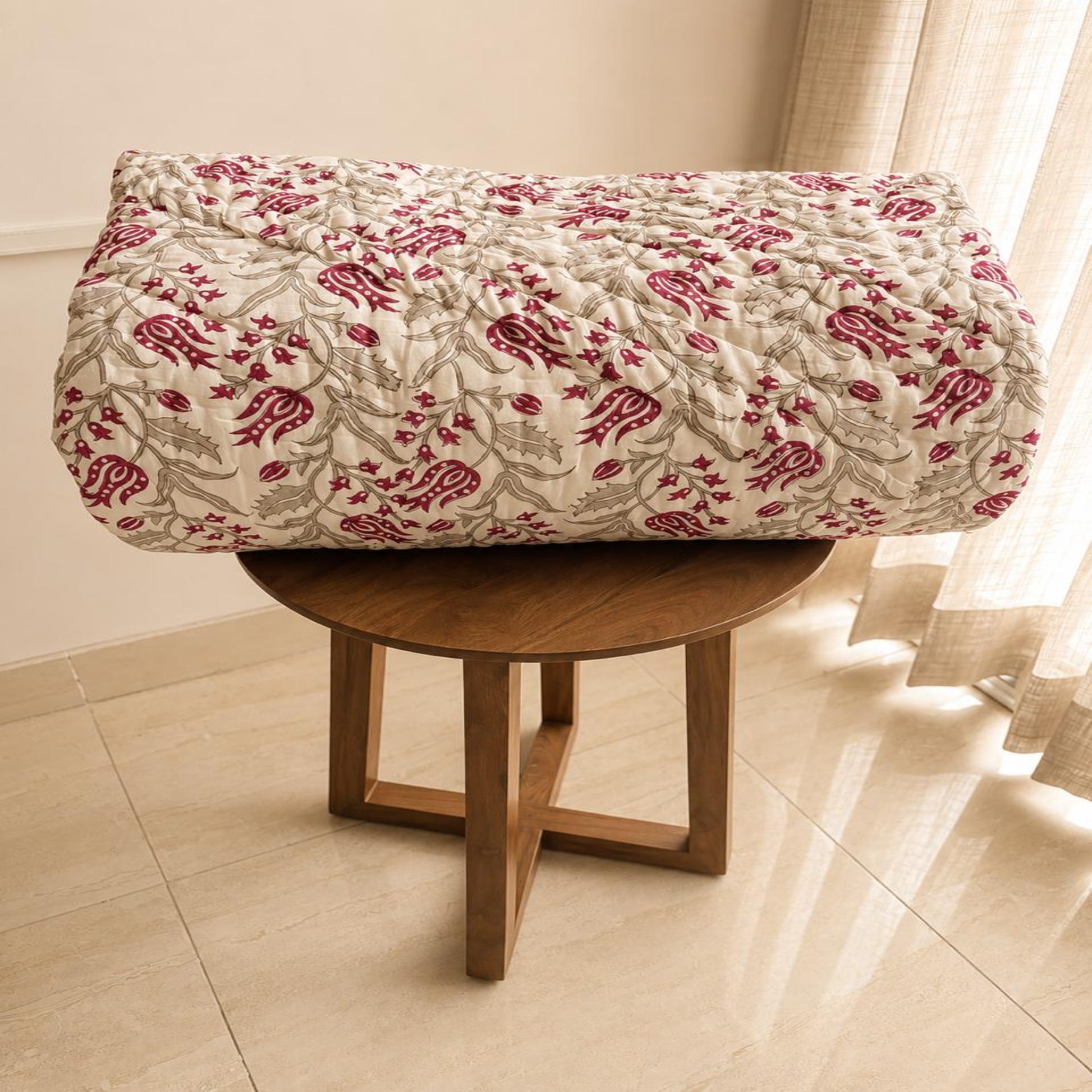 A folded Comforter/Razai/Quilt  with a red and gray floral pattern on a cream background, displayed on a round wooden stool in a brightly lit room.