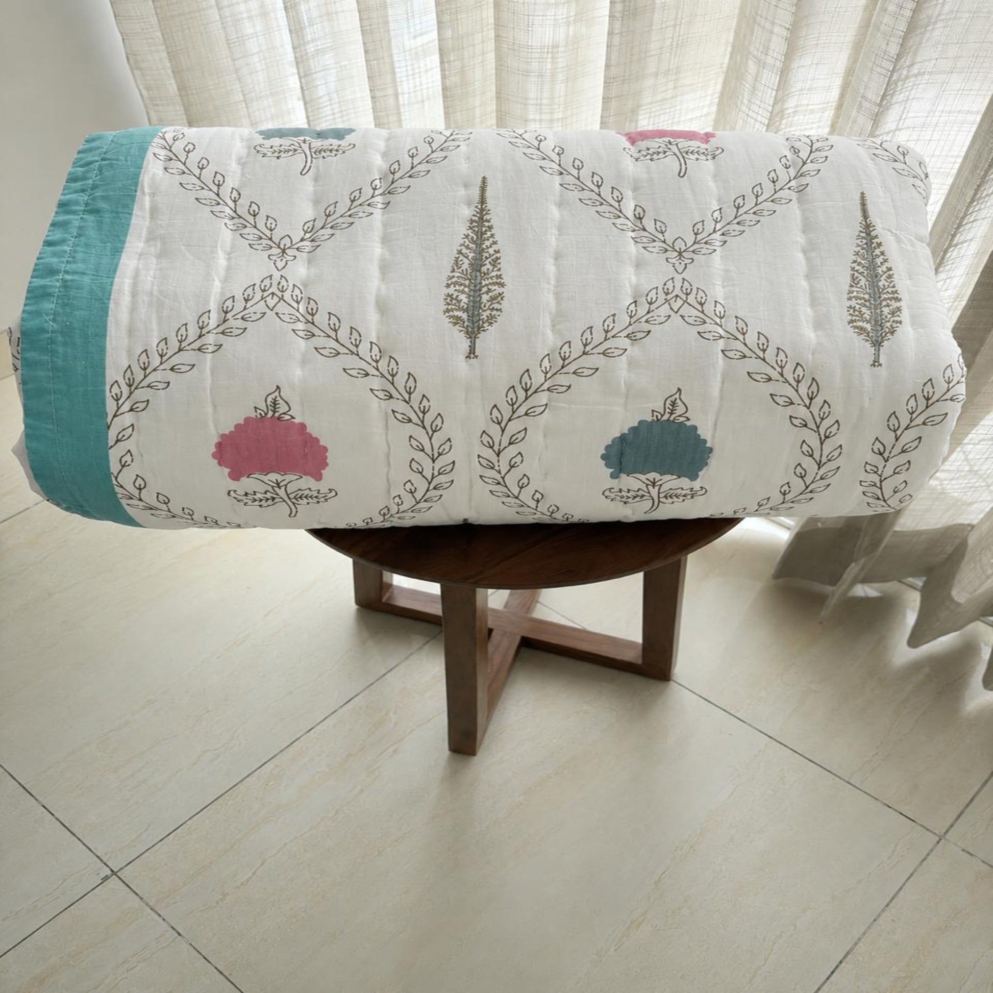A rolled multi coloured Jaipur Block Print Razai/Quilt/Comforter with a teal and gray botanical pattern and pink accents, displayed on a wooden stool in a bright room with sheer curtains.