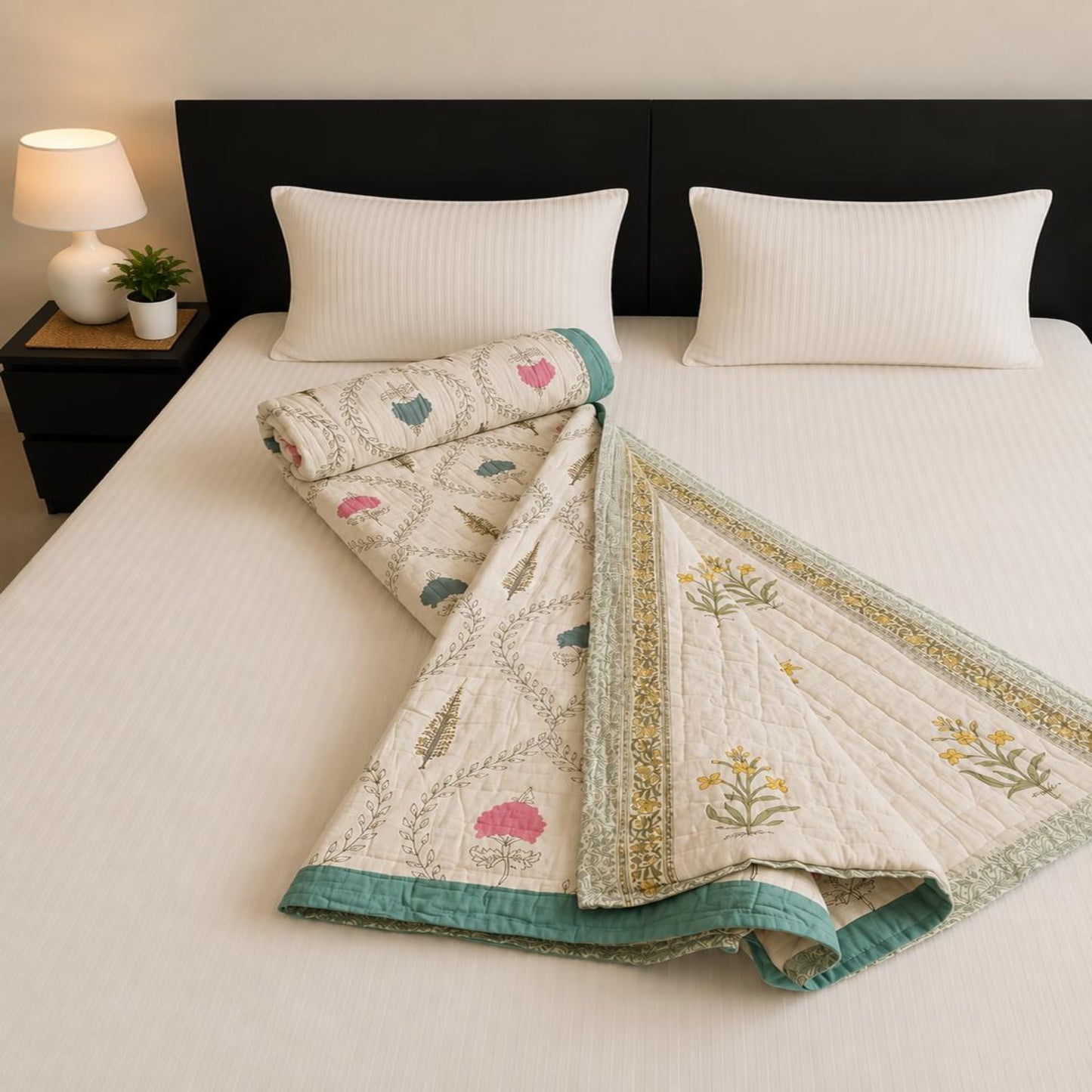 A folded and rolled Jaipuri Razai with floral design and teal borders, styled on a bed with white pillows and a dark headboard in a bedroom setting.