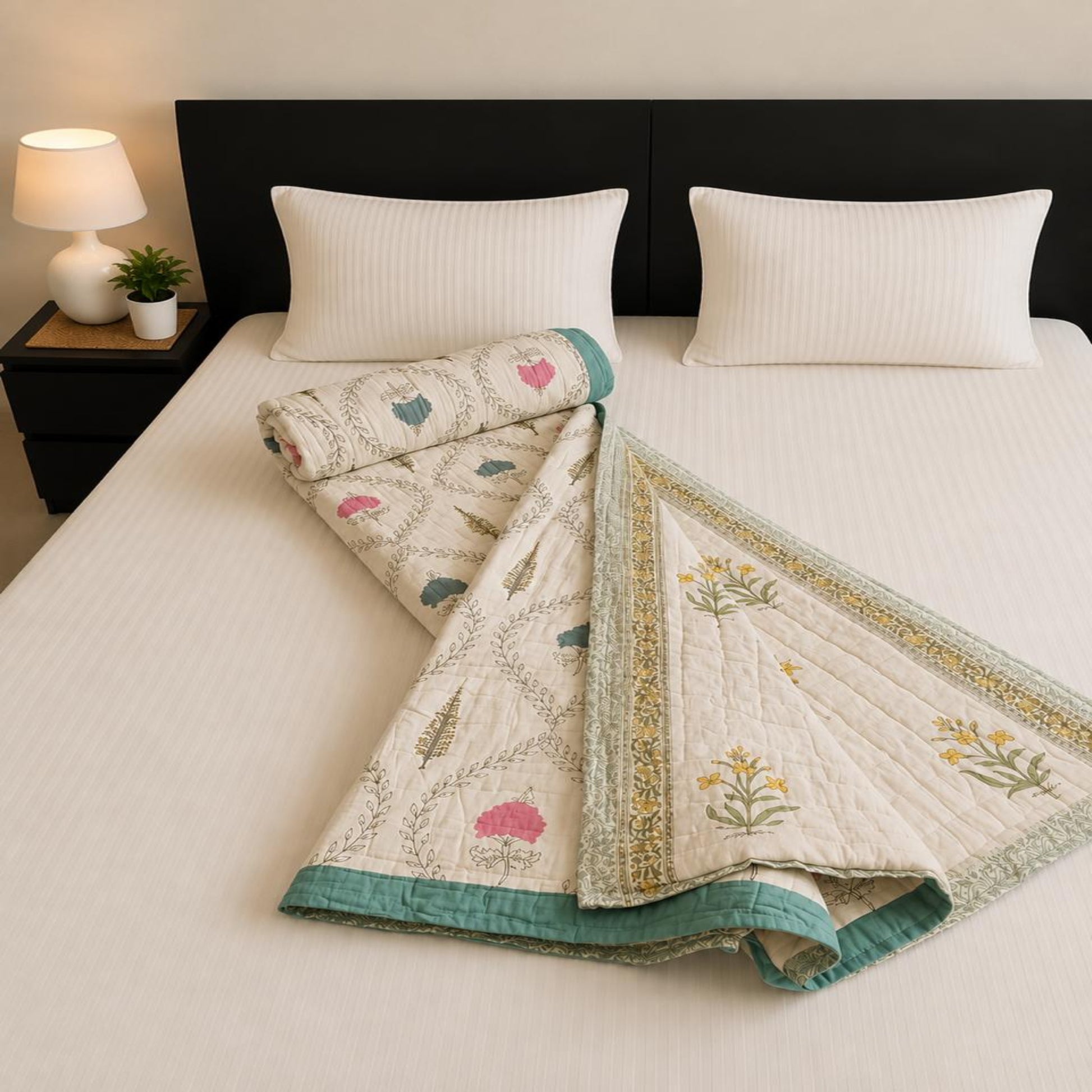 A folded and rolled Jaipuri Razai with floral design and teal borders, styled on a bed with white pillows and a dark headboard in a bedroom setting.