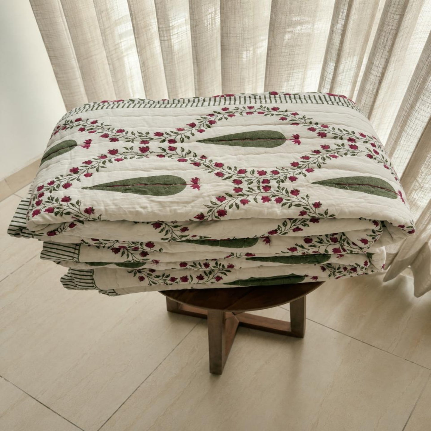 A folded Jaipuri Razai with a green and pink floral pattern on an off-white background, displayed on a wooden stool in front of sheer curtains.
