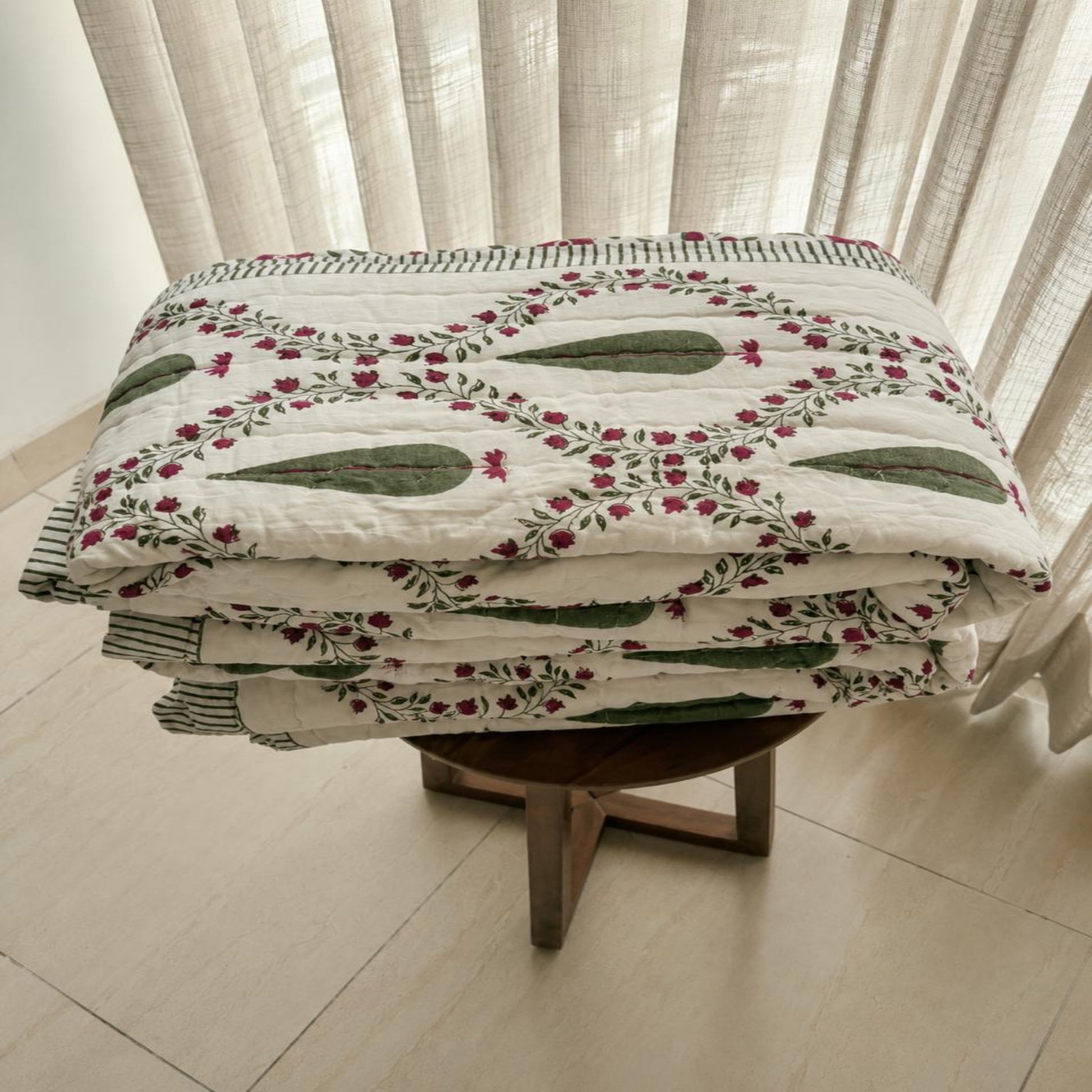 A folded Jaipuri Razai with a green and pink floral pattern on an off-white background, displayed on a wooden stool in front of sheer curtains.