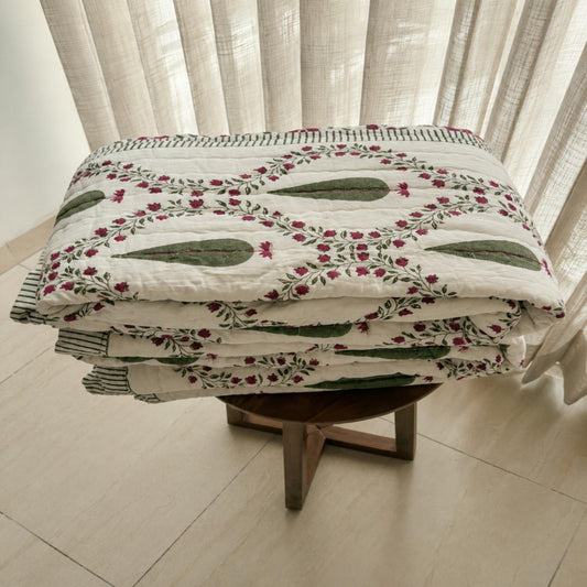 A folded Jaipuri Razai with a green and pink floral pattern on an off-white background, displayed on a wooden stool in front of sheer curtains.