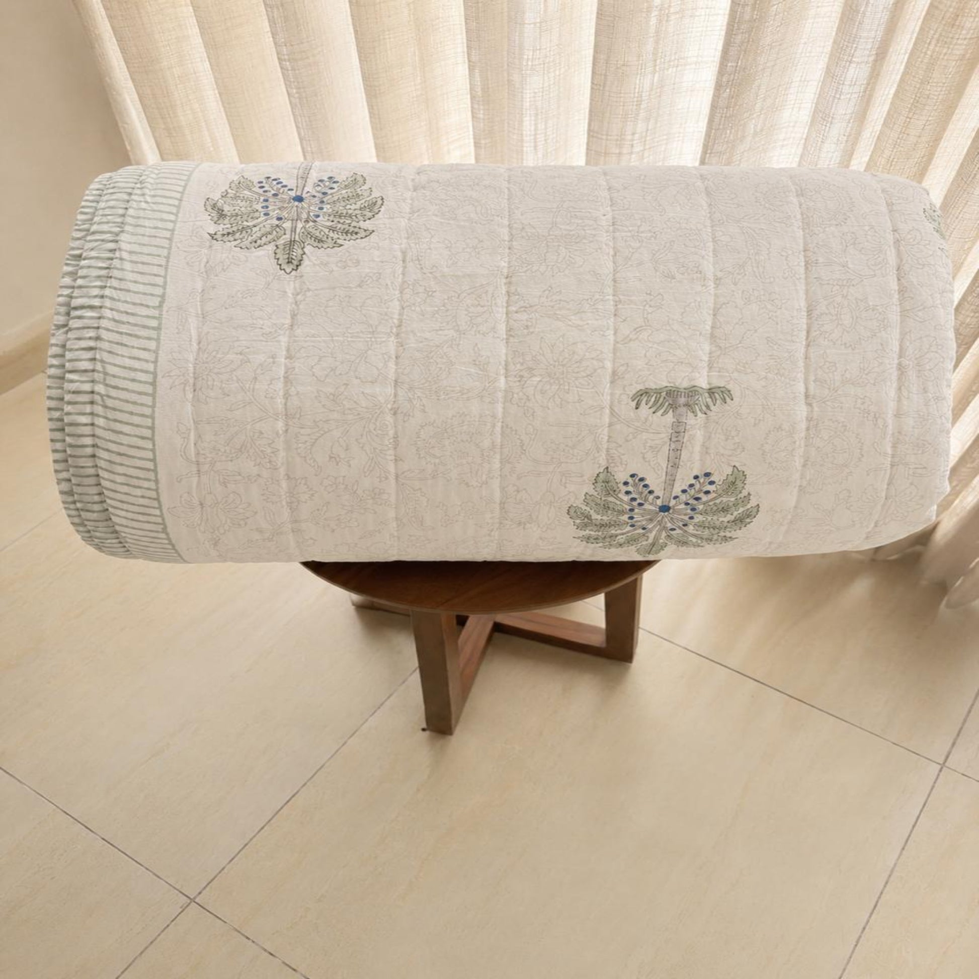A rolled Jaipuri Razai/Quilt/Comforter, displayed on a small wooden stool in front of sheer curtains. The fabric features vertical quilting, a ribbed knit edge, and two  floral motifs in muted green and blue.