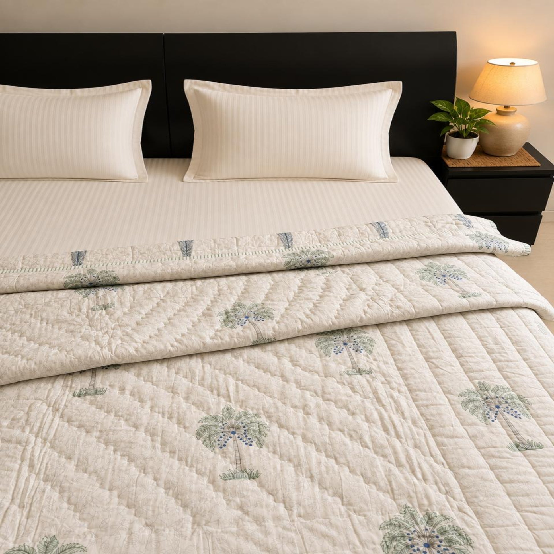 A neatly made bed with a folded beautiful Jaipuri Razai/Quilt/Comforter, featuring a subtle green palm tree pattern and matching pillow shams, styled in a bedroom setting with a dark headboard, bedside lamp, and small plant.