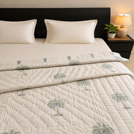 A neatly made bed with a folded beautiful Jaipuri Razai/Quilt/Comforter, featuring a subtle green palm tree pattern and matching pillow shams, styled in a bedroom setting with a dark headboard, bedside lamp, and small plant.