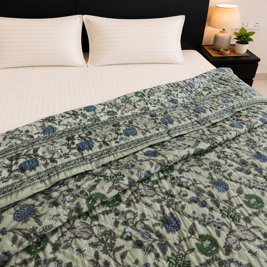 A floral patterned Jaipuri Quilt/Razai in sage green, blue, and olive tones is styled on a bed with white ribbed bedding and two pillows, next to a bedside lamp.