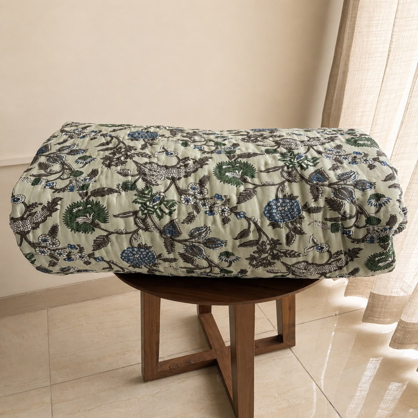 A beautiful Jaipuri Razai with a detailed botanical and floral pattern in green, blue, and gray on a light beige background, draped over a round wooden stool in an indoor setting.