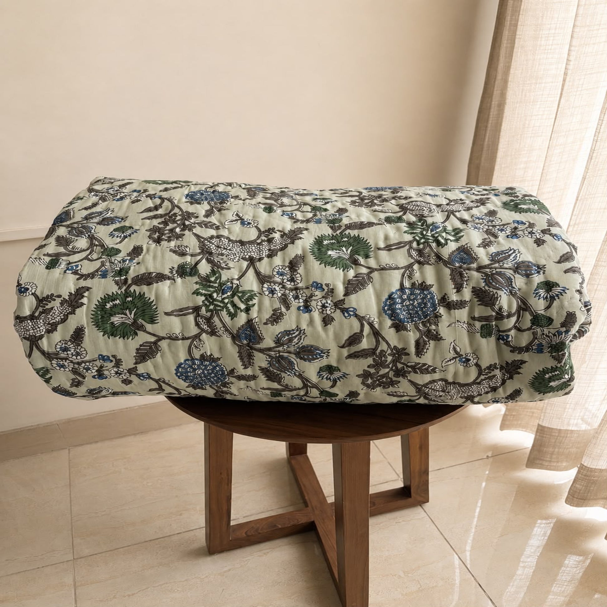 A beautiful Jaipuri Razai with a detailed botanical and floral pattern in green, blue, and gray on a light beige background, draped over a round wooden stool in an indoor setting.