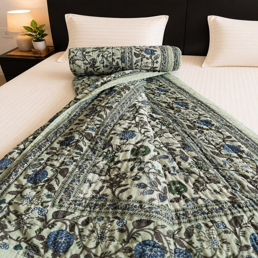 A beautiful Jaipuri Razai with blue and green floral motifs on an off-white and light sage green base, draped over a bed with white bedding and a rolled section showing the reverse side.