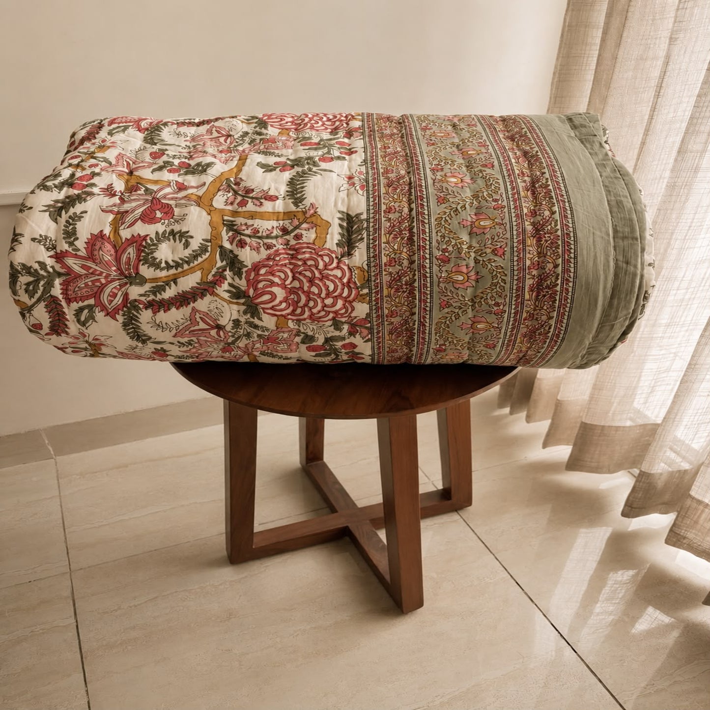 A rolled Jaipuri razai/quilt/comforter, displayed on a round wooden stool in an indoor room. The fabric features a cream base with a multicolor floral and botanical pattern in red, green, and mustard yellow, alongside a striped border.