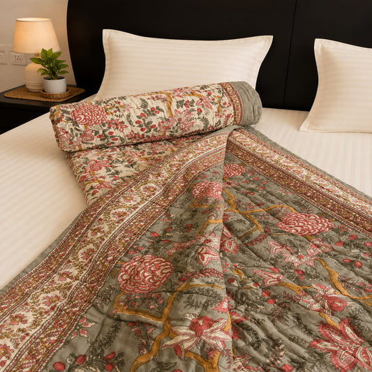 A Jaipuri Block Print Razai, a traditional floral and geometric pattern in muted olive green, red, and mustard yellow, styled on a bed with white pillows and a matching rolled-up quilt in a bedroom setting.