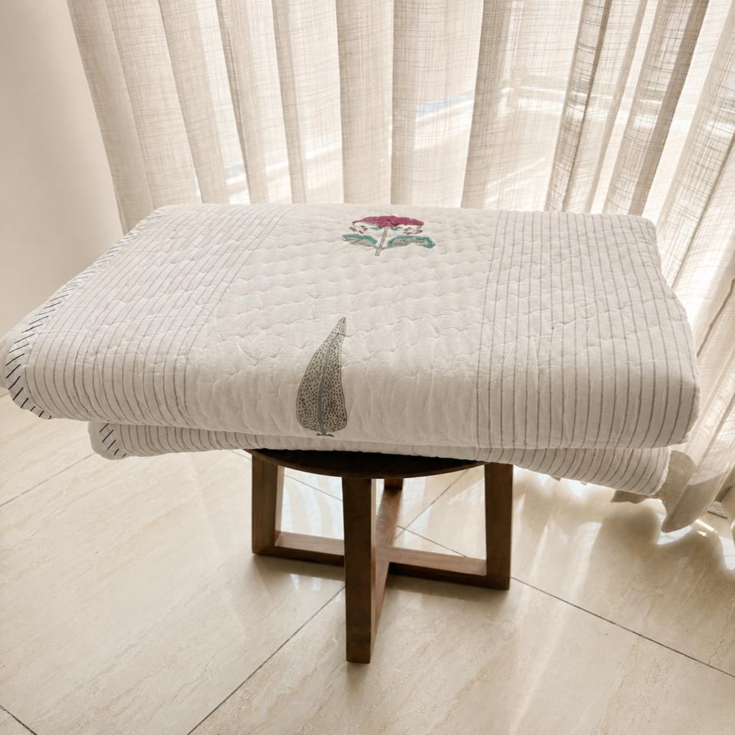 A folded beautiful Jaipuri Razai, displayed on a wooden stool in front of sheer white curtains.