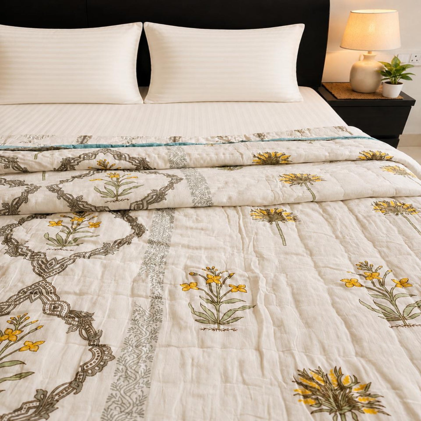 A close-up of a quilted bedspread with an off white base, featuring yellow floral bouquets and gray botanical chain motifs, styled on a bed with white pillows and a dark brown headboard.