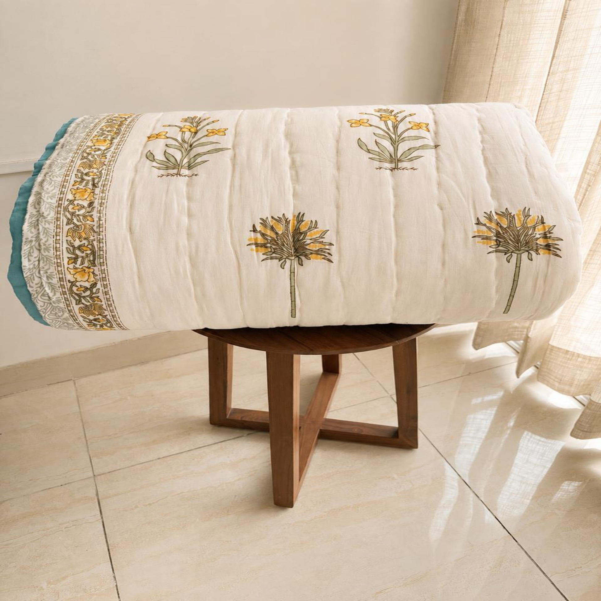 A beautiful block print comforter razai with yellow and green floral design and a decorative border, draped over a wooden stool in a bright room.