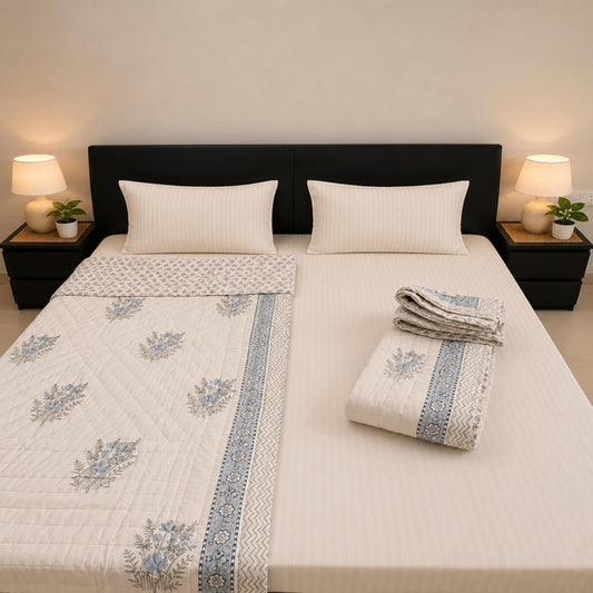 A neatly made bed with white bedding and a beautiful Cotton Block Print Jaipuri Razai on a white base featuring blue floral and border patterns, styled in a symmetric bedroom scene with two bedside tables, lamps, and plants.