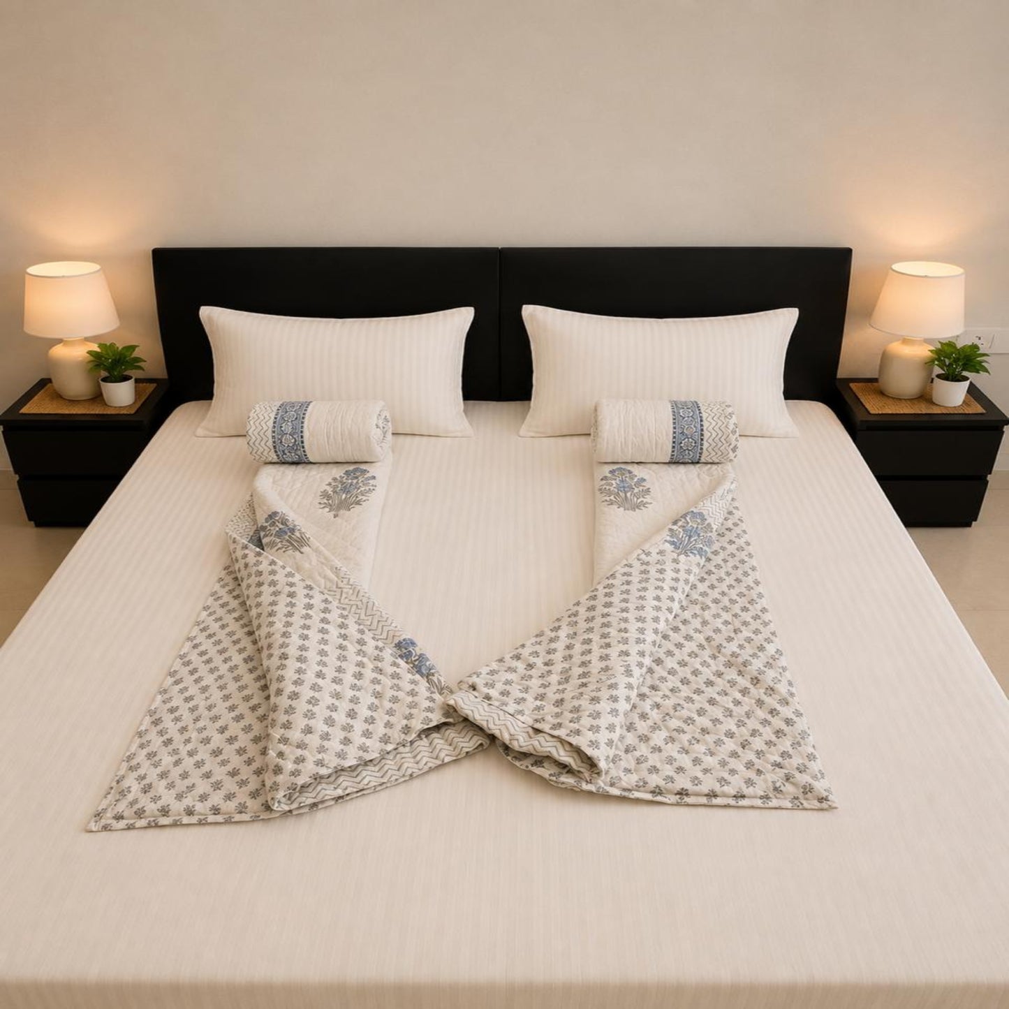 A neatly made bed with white bedding and two folded patterned Jaipuri Razai arranged symmetrically on either side of two matching rolled towels. The room has a dark headboard, bedside tables, lamps, and small plants, photographed from above.