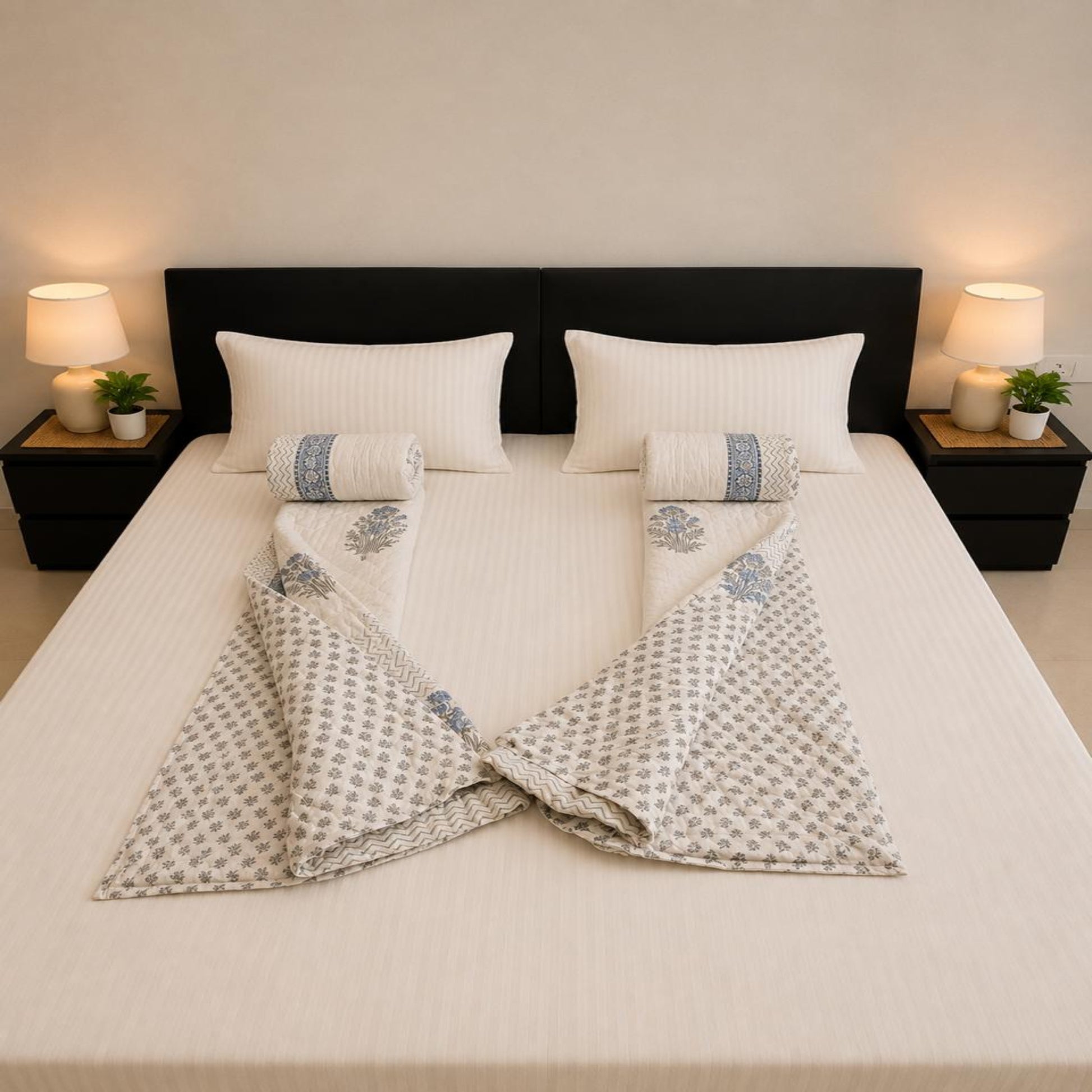 A neatly made bed with white bedding and two folded patterned Jaipuri Razai arranged symmetrically on either side of two matching rolled towels. The room has a dark headboard, bedside tables, lamps, and small plants, photographed from above.