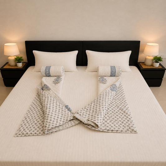 A neatly made bed with white bedding and two folded patterned Jaipuri Razai arranged symmetrically on either side of two matching rolled towels. The room has a dark headboard, bedside tables, lamps, and small plants, photographed from above.
