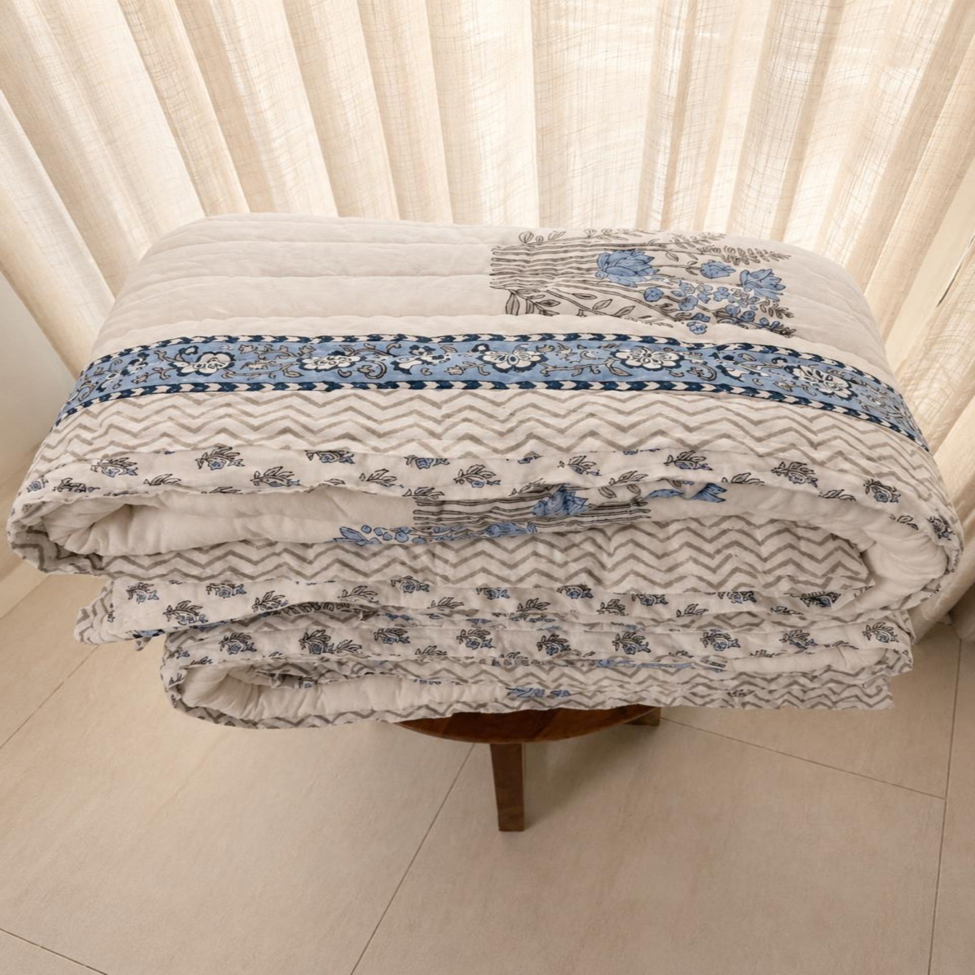 A folded Jaipuri Block Print Razai with a blue and gray patterned border and floral motifs, displayed on a small wooden stool in front of cream-colored curtains.